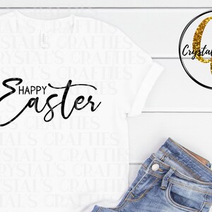 May include: White t-shirt with black text that reads "Happy Easter". The shirt is laying flat on a white wooden surface. A pair of blue jeans is laying on the surface to the right of the shirt. A gold glitter logo with the text "Crystal's Crafties" is in the upper right corner of the image.