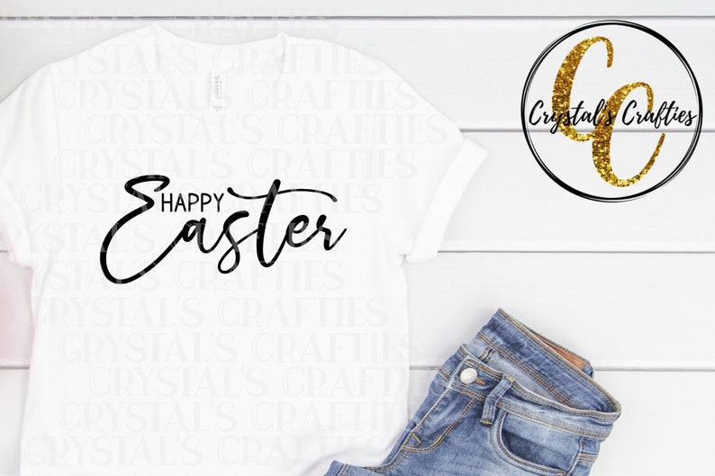 May include: White t-shirt with black text that reads "Happy Easter". The shirt is laying flat on a white wooden surface. A pair of blue jeans is laying on the surface to the right of the shirt. A gold glitter logo with the text "Crystal's Crafties" is in the upper right corner of the image.