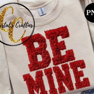 May include: A cream-colored sweatshirt with the words "BE MINE" in large, textured red letters. The sweatshirt also features a logo in a black circle with the text "Crystal's Crafties" and a gold glitter design. A black circle with "PNG" is in the upper right corner.