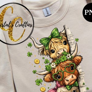 May include: A beige sweatshirt with a St. Patrick's Day theme. The design shows two cartoon Highland cows with shamrocks, gold coins, and green bows. The text "Crystal's Crafties" is in a gold glitter circle.