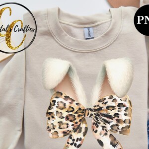 May include: A beige sweatshirt with a large leopard print bow with white fur accents. The bow is centered on the sweatshirt.