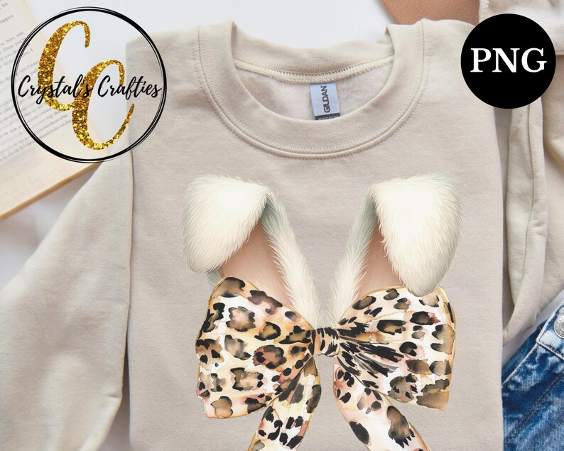 May include: A beige sweatshirt with a large leopard print bow with white fur accents. The bow is centered on the sweatshirt.
