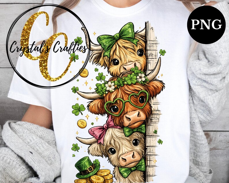 May include: White t-shirt with a colourful illustration of three Highland cows, each wearing St. Patrick's Day decorations. The cows have green bows, shamrock garlands, and heart-shaped glasses. Gold coins and shamrocks are scattered around.