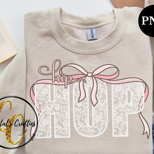 May include: A beige sweatshirt with a white lace design that spells out "HIP HOP" with a pink ribbon bow on top. The text "hip" is written in cursive.