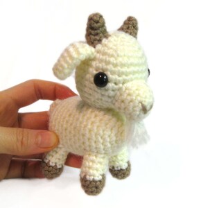May include: A white crocheted goat plush toy with brown horns and black eyes. The goat is standing on a white surface.