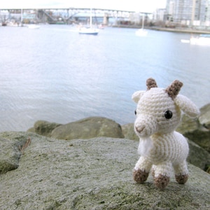May include: A white crocheted goat with brown ears and eyes. The goat is sitting on a gray rock.