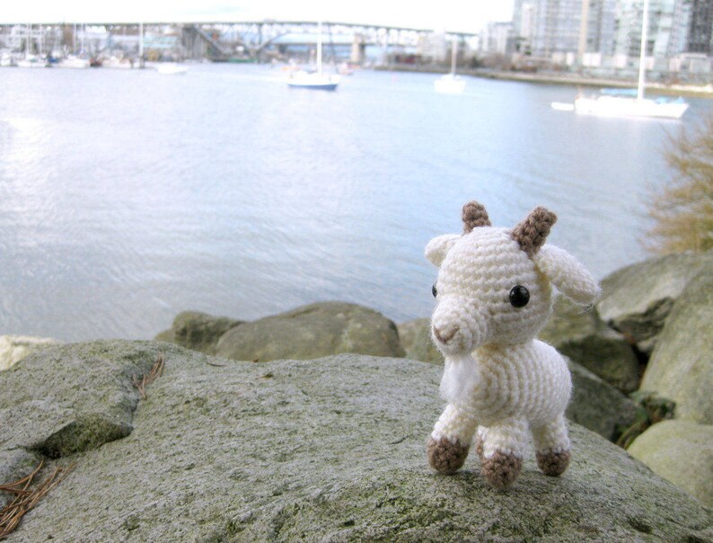 May include: A white crocheted goat with brown ears and eyes. The goat is sitting on a gray rock.