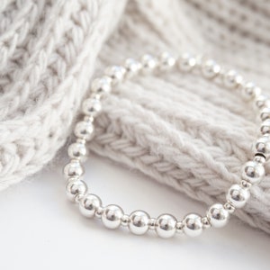 May include: A silver beaded bracelet with a simple design. The bracelet is made of round beads and is lying on a white knitted fabric.