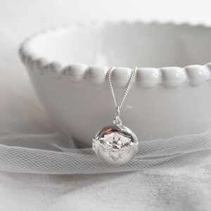 May include: A silver pendant necklace with a round locket, adorned with a bee motif. The locket hangs from a fine chain. The background features a white bowl and a sheer, white fabric.
