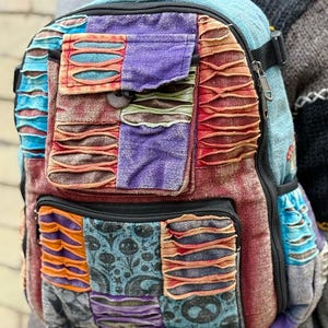 May include: A vibrant patchwork backpack with a mix of textures and designs. The backpack has several pockets, a zip closure, and adjustable straps. The colour scheme includes blue, purple, orange, and red.