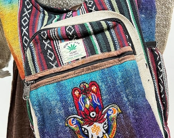 May include: A colorful hemp shoulder bag with a front pocket and a zipper closure. The bag features a vibrant embroidered Hamsa hand design on a blue and purple gradient background. The bag has a striped pattern in various colors.