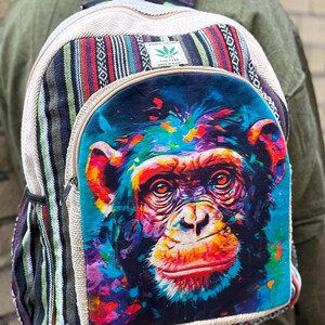 May include: A colourful backpack with a vibrant chimpanzee portrait on the front. The backpack has a beige, striped, and patterned design with a zip closure. The image includes a small green leaf logo.