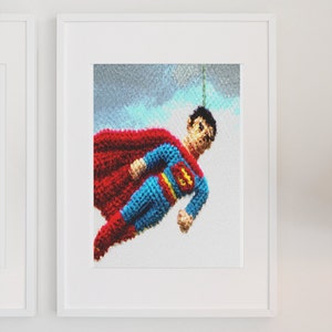 Può includere: Una figura di Superman all'uncinetto con un mantello rosso e un costume blu vola su uno sfondo blu e bianco. La figura è incorniciata in una cornice bianca.