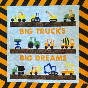 May include: A colorful quilt featuring various construction vehicles in yellow, blue, green, and orange. The words "BIG TRUCKS" and "BIG DREAMS" are displayed in orange block letters. The quilt has a light blue background and a black and yellow striped border.