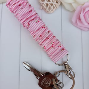 Pode incluir: Um porta-chaves de tecido listrado rosa e branco com um padr&atilde;o de cerejas. O porta-chaves &eacute; preso a um porta-chaves e fecho prateados, com uma chave. O tecido &eacute; franzido.