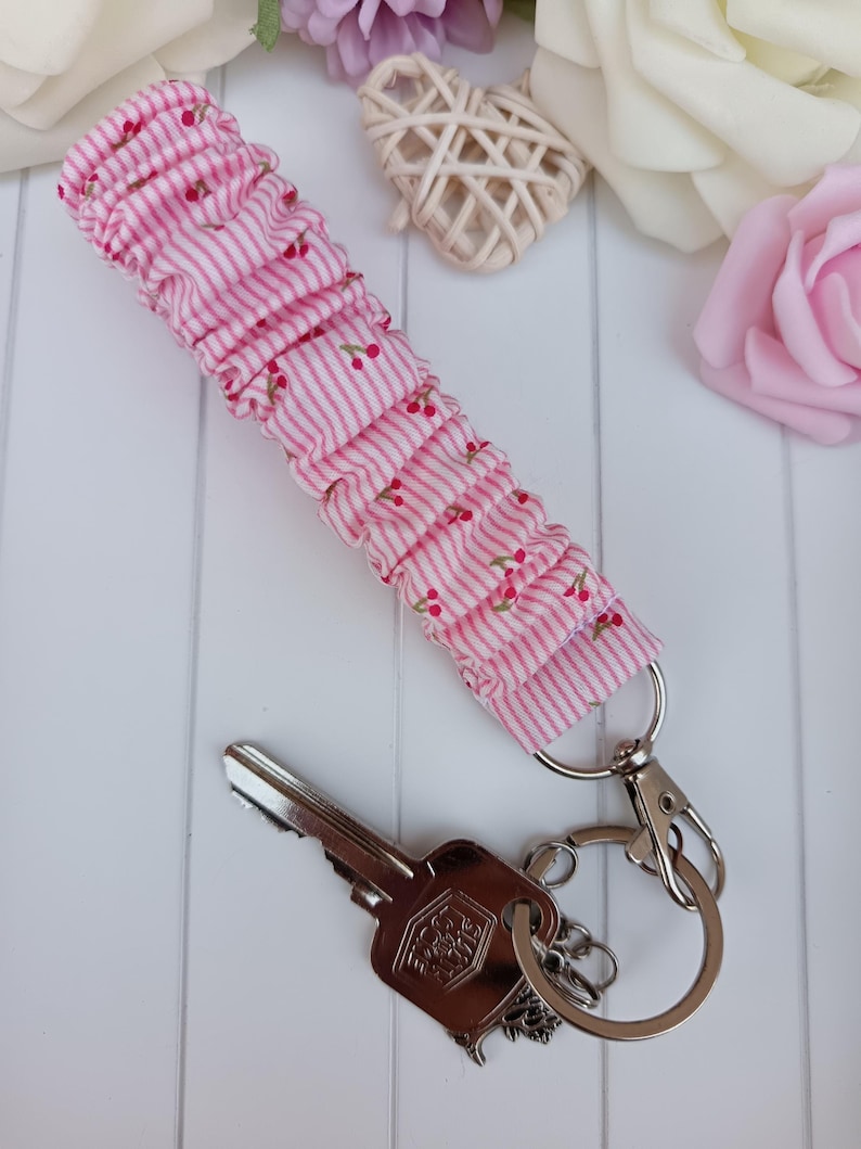 Pode incluir: Um porta-chaves de tecido listrado rosa e branco com um padr&atilde;o de cerejas. O porta-chaves &eacute; preso a um porta-chaves e fecho prateados, com uma chave. O tecido &eacute; franzido.