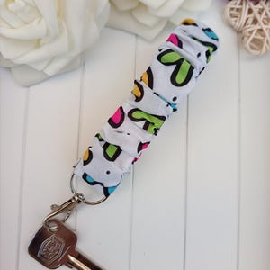 Pode incluir: Um porta-chaves de tecido branco com um padr&atilde;o colorido de formas verdes, rosa e azuis. O porta-chaves est&aacute; preso a um mosquet&atilde;o prateado e a uma chave. O fundo inclui rosas brancas e uma superf&iacute;cie de madeira branca.