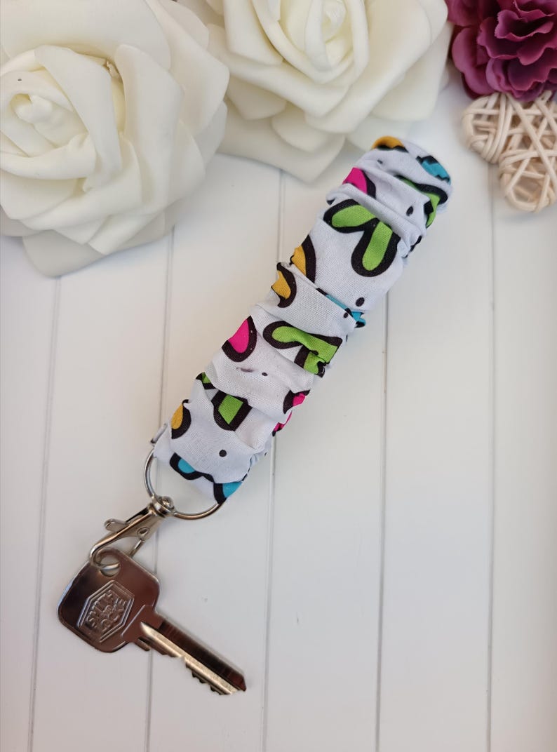 Pode incluir: Um porta-chaves de tecido branco com um padr&atilde;o colorido de formas verdes, rosa e azuis. O porta-chaves est&aacute; preso a um mosquet&atilde;o prateado e a uma chave. O fundo inclui rosas brancas e uma superf&iacute;cie de madeira branca.