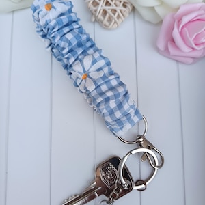 Pode incluir: Uma pulseira de chaveiro azul e branca com detalhes de margaridas brancas e um fecho prateado. A pulseira est&aacute; presa a um conjunto de chaves com um pingente de &aacute;rvore.