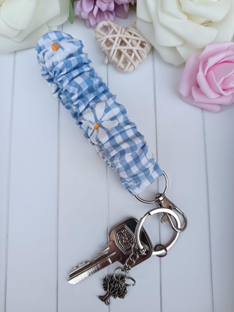 Pode incluir: Uma pulseira de chaveiro azul e branca com detalhes de margaridas brancas e um fecho prateado. A pulseira est&aacute; presa a um conjunto de chaves com um pingente de &aacute;rvore.