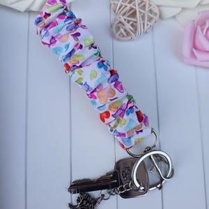 Pode incluir: Um porta-chaves de tecido branco com um padr&atilde;o de cora&ccedil;&atilde;o colorido, preso a um porta-chaves prateado com duas chaves. Uma chave tem um pingente de &aacute;rvore. A pulseira &eacute; enrugada e tem cora&ccedil;&otilde;es rosa, roxos, azuis e amarelos.