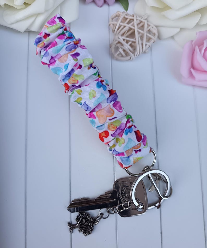 Pode incluir: Um porta-chaves de tecido branco com um padr&atilde;o de cora&ccedil;&atilde;o colorido, preso a um porta-chaves prateado com duas chaves. Uma chave tem um pingente de &aacute;rvore. A pulseira &eacute; enrugada e tem cora&ccedil;&otilde;es rosa, roxos, azuis e amarelos.