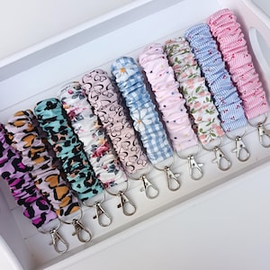 Pode incluir: Uma cole&ccedil;&atilde;o de chaveiros de pulso de tecido em v&aacute;rios padr&otilde;es e cores, incluindo estampa de leopardo, floral e xadrez. Cada chaveiro tem um fecho prateado para prender as chaves.