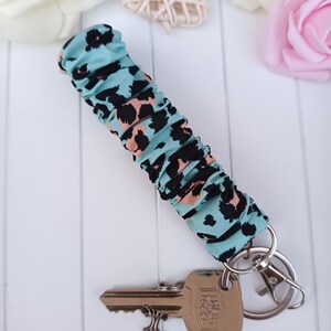Pode incluir: Um porta-chaves com tecido estampado de leopardo turquesa. O tecido &eacute; franzido e preso a um porta-chaves e mosquet&atilde;o prateados. Uma chave est&aacute; presa. No fundo, flores brancas e rosas.
