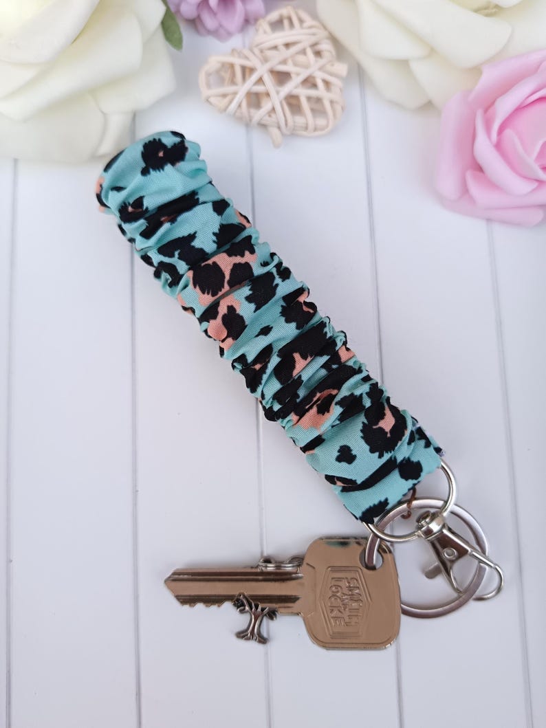 Pode incluir: Um porta-chaves com tecido estampado de leopardo turquesa. O tecido &eacute; franzido e preso a um porta-chaves e mosquet&atilde;o prateados. Uma chave est&aacute; presa. No fundo, flores brancas e rosas.