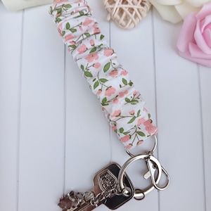 Pode incluir: Um porta-chaves de tecido com fundo branco e padr&atilde;o floral rosa e folhas verdes. Est&aacute; preso a um porta-chaves prateado e a uma chave. O tecido &eacute; franzido. A chave tem um berloque decorativo.