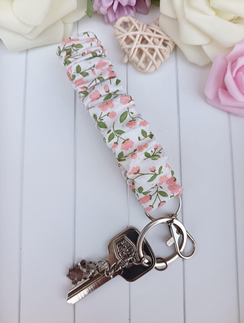 Pode incluir: Um porta-chaves de tecido com fundo branco e padr&atilde;o floral rosa e folhas verdes. Est&aacute; preso a um porta-chaves prateado e a uma chave. O tecido &eacute; franzido. A chave tem um berloque decorativo.