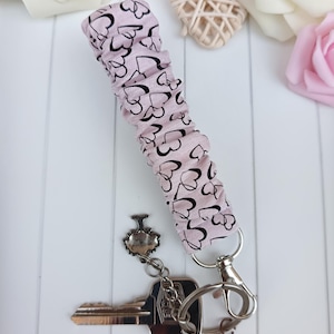 Pode incluir: Uma pulseira de chave rosa claro com um padr&atilde;o de cora&ccedil;&atilde;o preto. A pulseira est&aacute; presa a um porta-chaves prateado e a uma chave. O fundo inclui flores brancas e rosas.