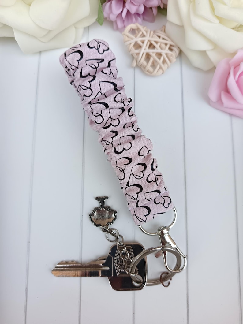 Pode incluir: Uma pulseira de chave rosa claro com um padr&atilde;o de cora&ccedil;&atilde;o preto. A pulseira est&aacute; presa a um porta-chaves prateado e a uma chave. O fundo inclui flores brancas e rosas.
