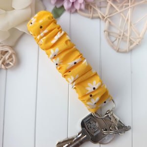 Pode incluir: Um porta-chaves de tecido amarelo com um padr&atilde;o de margaridas brancas. V&aacute;rias chaves est&atilde;o presas a um porta-chaves prateado. O tecido &eacute; franzido, criando uma apar&ecirc;ncia texturizada. O fundo inclui flores e uma superf&iacute;cie branca.
