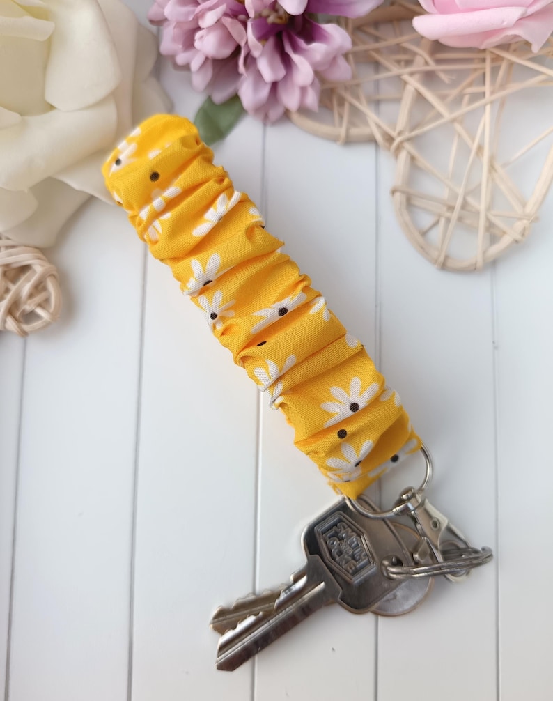 Pode incluir: Um porta-chaves de tecido amarelo com um padr&atilde;o de margaridas brancas. V&aacute;rias chaves est&atilde;o presas a um porta-chaves prateado. O tecido &eacute; franzido, criando uma apar&ecirc;ncia texturizada. O fundo inclui flores e uma superf&iacute;cie branca.