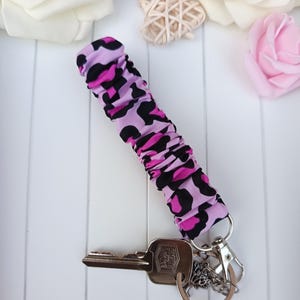 Pode incluir: Um porta-chaves com um tecido com estampa de leopardo rosa e preto. O porta-chaves &eacute; preso a um porta-chaves prateado com chaves e um pingente decorativo. O fundo inclui rosas brancas e uma decora&ccedil;&atilde;o em forma de cora&ccedil;&atilde;o.