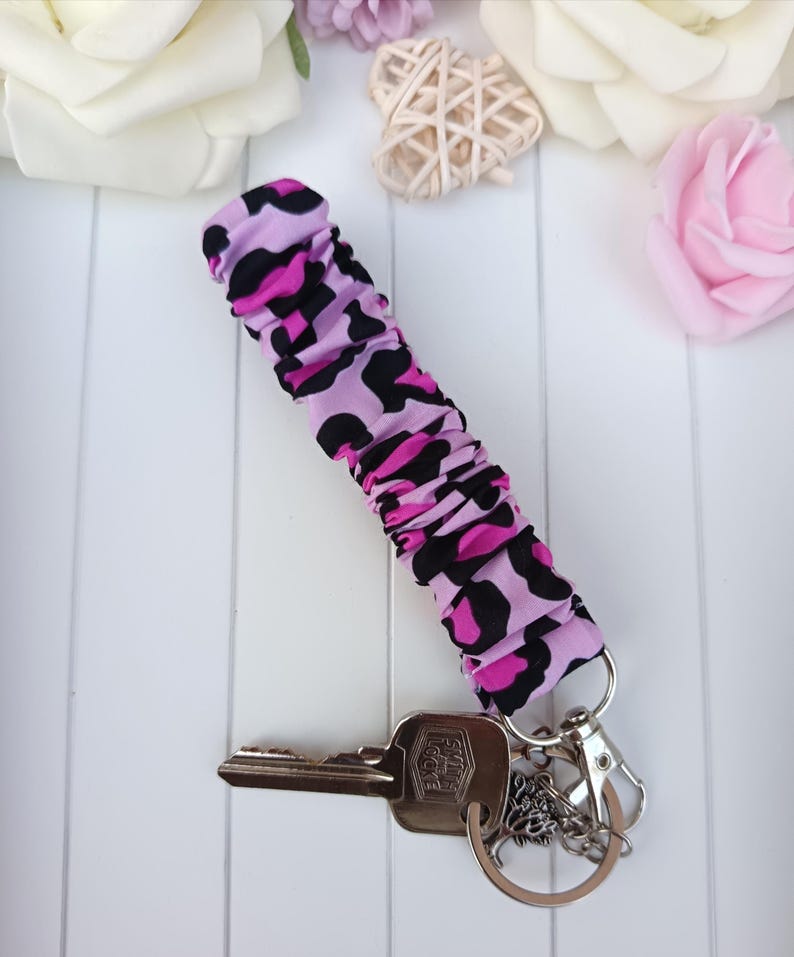 Pode incluir: Um porta-chaves com um tecido com estampa de leopardo rosa e preto. O porta-chaves &eacute; preso a um porta-chaves prateado com chaves e um pingente decorativo. O fundo inclui rosas brancas e uma decora&ccedil;&atilde;o em forma de cora&ccedil;&atilde;o.