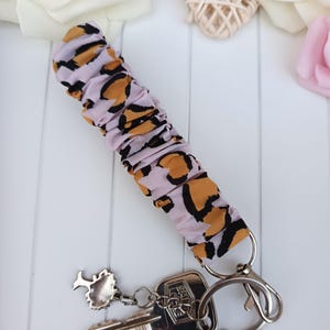 Pode incluir: Uma pulseira de chaveiro com tecido estampado de leopardo lil&aacute;s claro, um fecho prateado e um porta-chaves. O porta-chaves cont&eacute;m duas chaves e um pequeno pingente de &aacute;rvore. A pulseira tem cerca de 15 cm de comprimento.