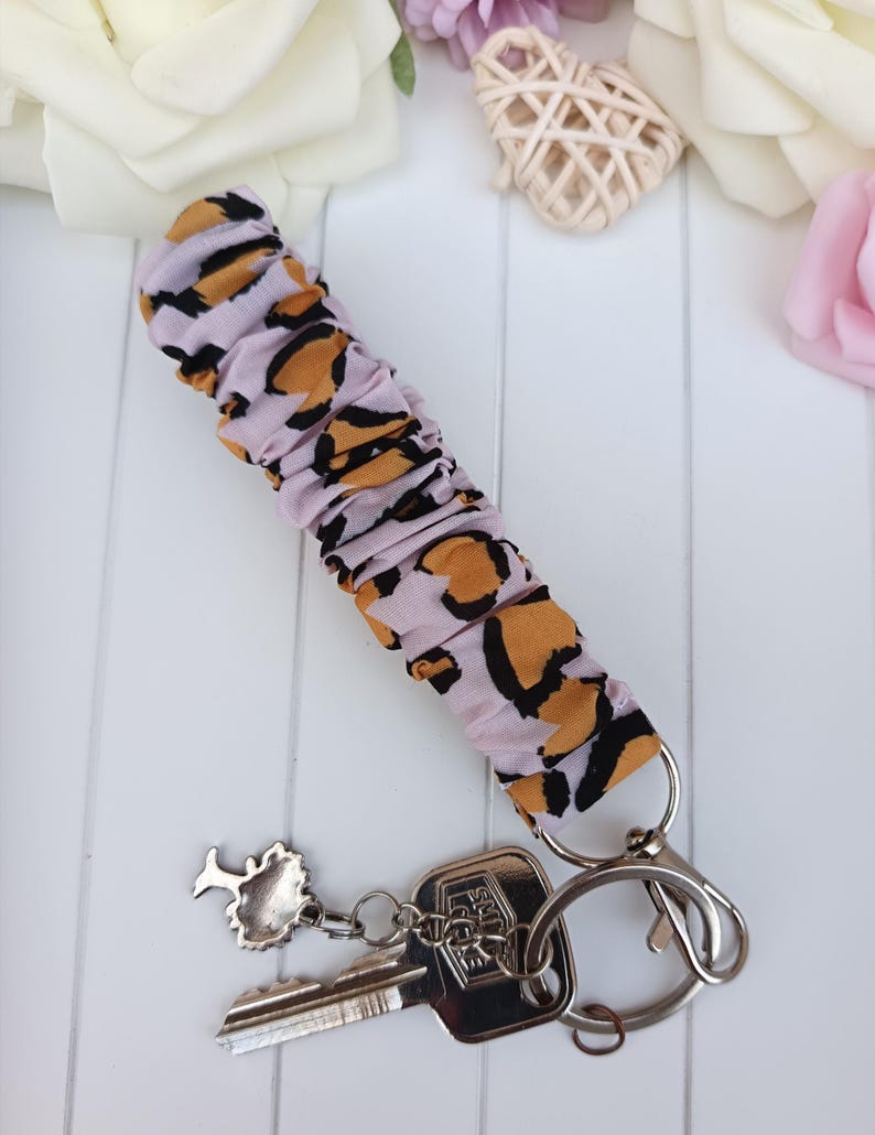 Pode incluir: Uma pulseira de chaveiro com tecido estampado de leopardo lil&aacute;s claro, um fecho prateado e um porta-chaves. O porta-chaves cont&eacute;m duas chaves e um pequeno pingente de &aacute;rvore. A pulseira tem cerca de 15 cm de comprimento.