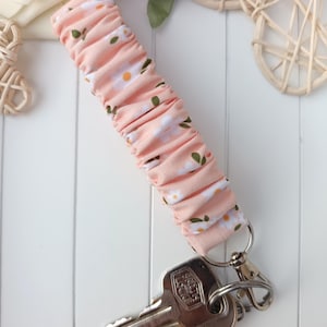 Pode incluir: Uma pulseira de chaveiro de tecido rosa com um padr&atilde;o de margaridas brancas, presa a um chaveiro prateado e a um conjunto de chaves. A pulseira &eacute; feita de tecido franzido.