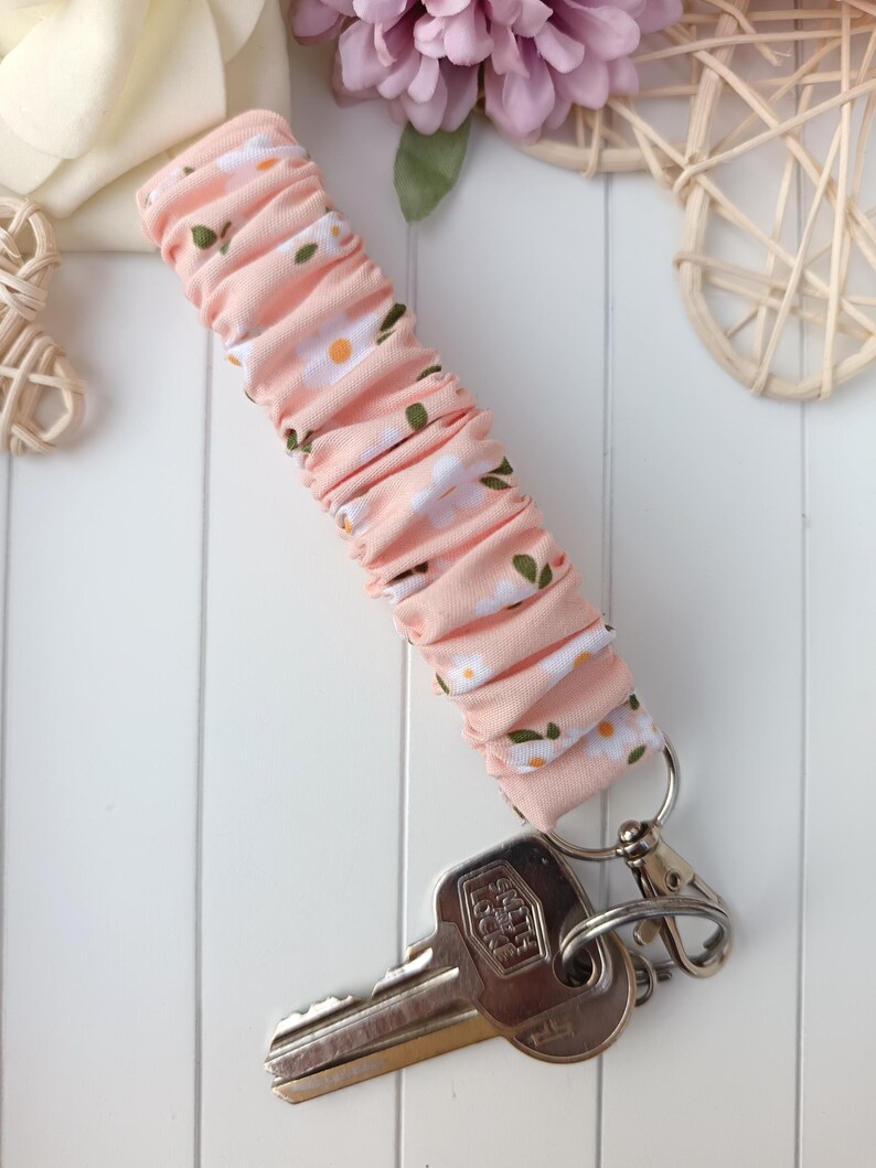 Pode incluir: Uma pulseira de chaveiro de tecido rosa com um padr&atilde;o de margaridas brancas, presa a um chaveiro prateado e a um conjunto de chaves. A pulseira &eacute; feita de tecido franzido.