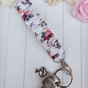 Pode incluir: Uma pulseira de chave floral branca com fecho e chaveiro prateados. O tecido apresenta um padr&atilde;o de rosas rosa, vermelhas e amarelas com folhas verdes. Uma chave e um pequeno encanto est&atilde;o anexados.