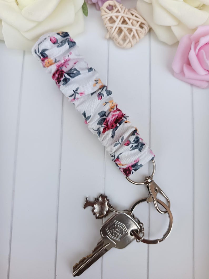 Pode incluir: Uma pulseira de chave floral branca com fecho e chaveiro prateados. O tecido apresenta um padr&atilde;o de rosas rosa, vermelhas e amarelas com folhas verdes. Uma chave e um pequeno encanto est&atilde;o anexados.