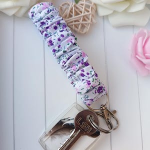 Pode incluir: Um porta-chaves de pulso em tecido floral com um porta-chaves em acr&iacute;lico transparente. A pulseira apresenta um fundo branco com estampas de flores roxas e rosas. O porta-chaves tem um desenho de pena e uma chave com as letras "LSH".