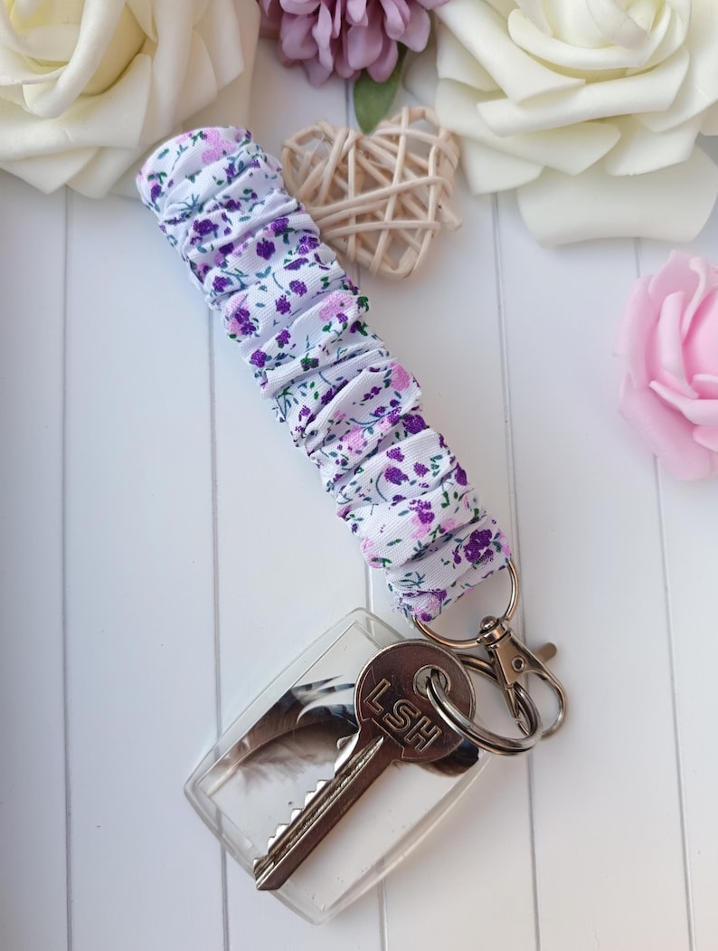 Pode incluir: Um porta-chaves de pulso em tecido floral com um porta-chaves em acr&iacute;lico transparente. A pulseira apresenta um fundo branco com estampas de flores roxas e rosas. O porta-chaves tem um desenho de pena e uma chave com as letras "LSH".