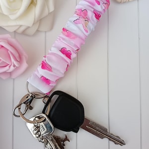 Pode incluir: Um porta-chaves de pulso com estampa de borboletas rosa e branco, argolas de chaveiro prateadas e chaves do carro. A pulseira de tecido &eacute; franzida e apresenta um padr&atilde;o repetido de borboletas rosa. As chaves s&atilde;o prateadas e pretas.