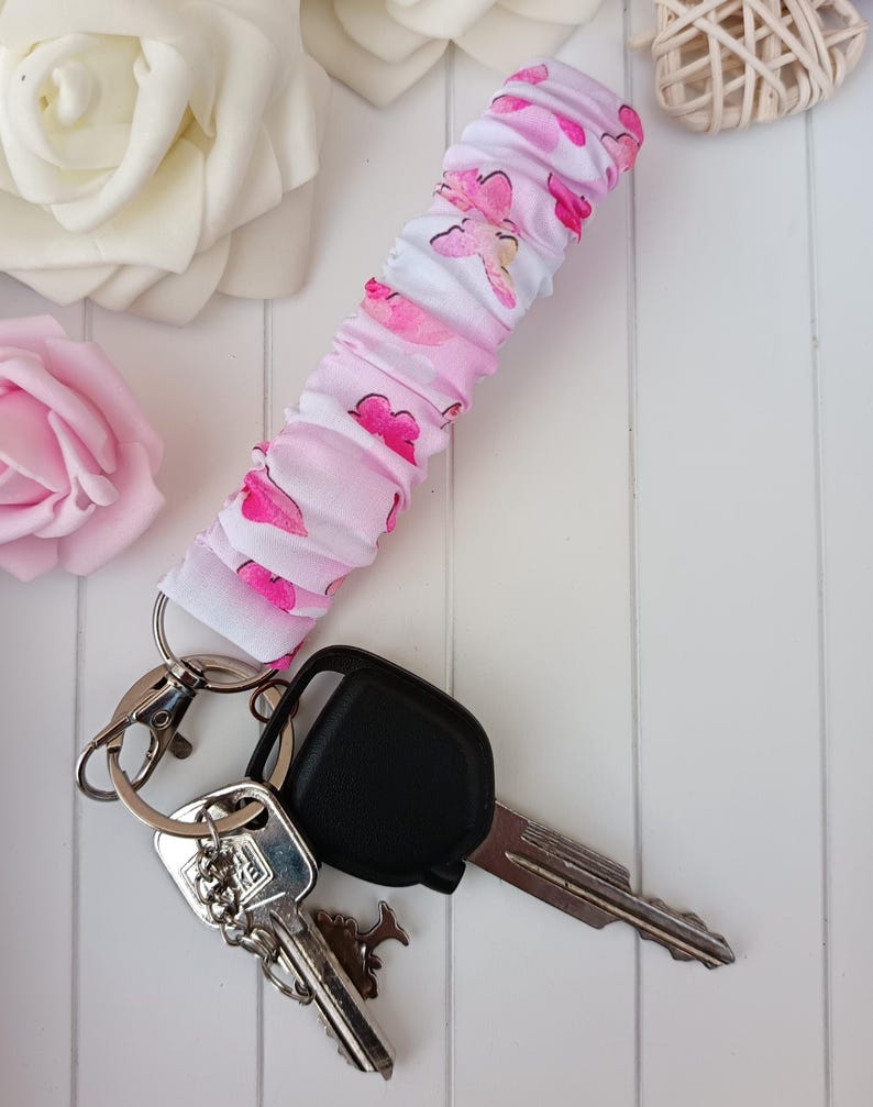 Pode incluir: Um porta-chaves de pulso com estampa de borboletas rosa e branco, argolas de chaveiro prateadas e chaves do carro. A pulseira de tecido &eacute; franzida e apresenta um padr&atilde;o repetido de borboletas rosa. As chaves s&atilde;o prateadas e pretas.