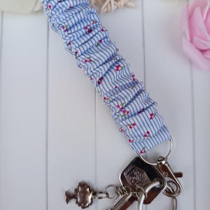 Pode incluir: Um porta-chaves de pulso de tecido listrado azul e branco com pequenos detalhes florais vermelhos. O porta-chaves &eacute; preso a um porta-chaves prateado com chaves e um pingente decorativo.