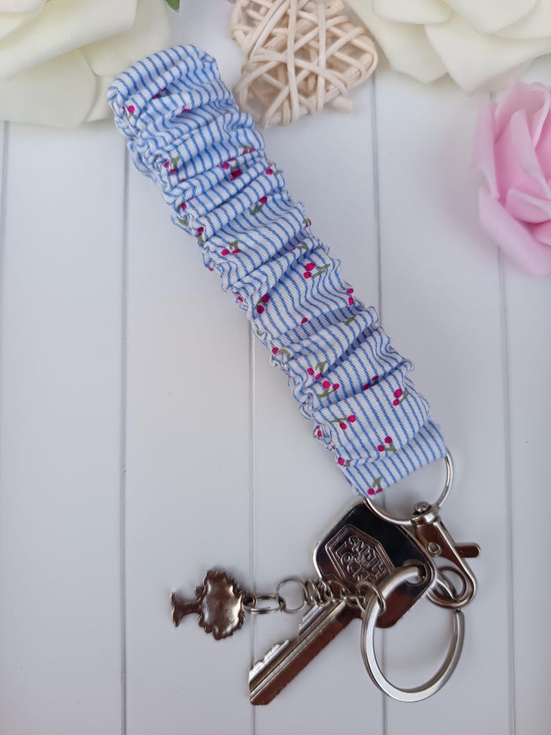 Pode incluir: Um porta-chaves de pulso de tecido listrado azul e branco com pequenos detalhes florais vermelhos. O porta-chaves &eacute; preso a um porta-chaves prateado com chaves e um pingente decorativo.