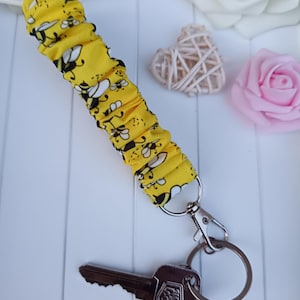 Pode incluir: Porta-chaves de tecido amarelo com um padr&atilde;o de abelhas, preso a um porta-chaves e fecho prateados. Uma chave com um pendente decorativo tamb&eacute;m est&aacute; presa. O fundo inclui flores decorativas e um objeto em forma de cora&ccedil;&atilde;o tecido.